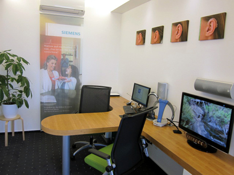 Modernes Hörakustiker-Büro mit Schreibtisch, Computer, Ohrbildern und Siemens-Plakat.